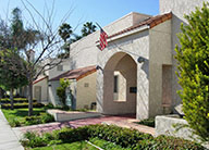 Exterior of a building at Coral Wood Court, with a red tile path to an archway to mission-style building in beige stucco with a red tile roof.